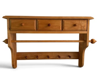 Peut inclure: Une étagère murale en bois brun clair comprenant trois petits tiroirs avec des boutons ronds en haut, une tige horizontale pour suspendre des objets au milieu et une barre inférieure équipée de cinq petites patères pour le rangement.