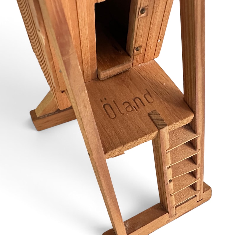 May include: A wooden model of a windmill structure featuring a vertical main body with a small doorway, a platform base with the word &Ouml;land embossed into the wood, and a side ladder attached to the support frame, all crafted from light brown wood.