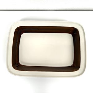 Puede incluir: Una fuente de horno rectangular de cerámica con un centro liso de color crema y un borde ancho esmaltado en marrón oscuro. El borde exterior de la fuente presenta un patrón texturizado sutil que recorre todo el perímetro del borde.