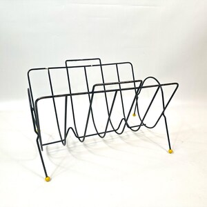 May include: Black metal magazine rack with a mid-century modern design. The rack features a series of vertical and curved wire supports, with yellow ball feet. The rack is designed to hold magazines or other reading materials.