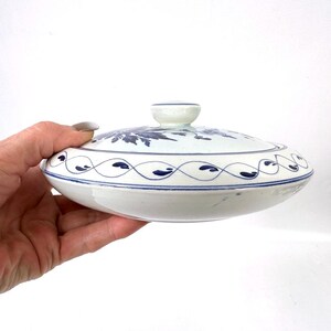 May include: A white ceramic dish with a lid, decorated with blue floral and wave patterns. The lid has a small, round handle. The dish is held in a hand against a white background. The dish is approximately 15 cm in diameter.