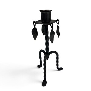 May include: A black metal candlestick holder featuring a central twisted stem supported by three curved, twisted legs. The top platform holds a cylindrical candle cup and is decorated with four hanging metal leaf-shaped ornaments at the corners.
