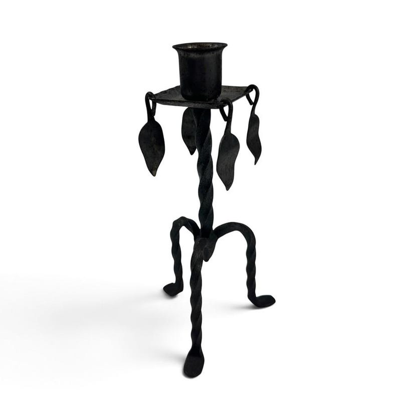 May include: A black metal candlestick holder featuring a central twisted stem supported by three curved, twisted legs. The top platform holds a cylindrical candle cup and is decorated with four hanging metal leaf-shaped ornaments at the corners.