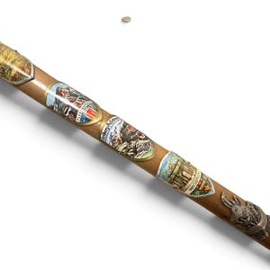 Puede incluir: Un bast&oacute;n de madera de color marr&oacute;n claro decorado con varios escudos met&aacute;licos de recuerdo. Los escudos presentan ilustraciones coloridas de paisajes monta&ntilde;osos y edificios, con texto que incluye Flexenstrasse, Arlberg-Passstrasse e Innsbruck.