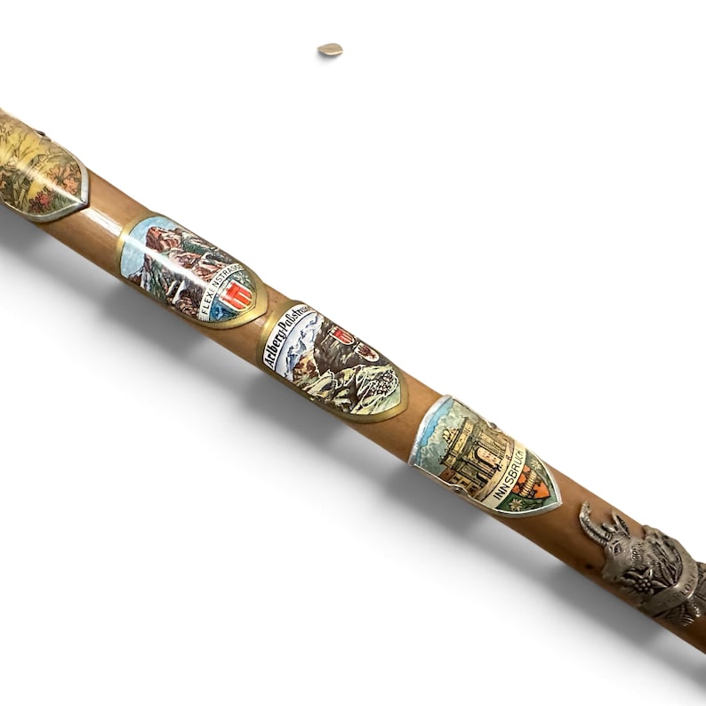 Puede incluir: Un bast&oacute;n de madera de color marr&oacute;n claro decorado con varios escudos met&aacute;licos de recuerdo. Los escudos presentan ilustraciones coloridas de paisajes monta&ntilde;osos y edificios, con texto que incluye Flexenstrasse, Arlberg-Passstrasse e Innsbruck.