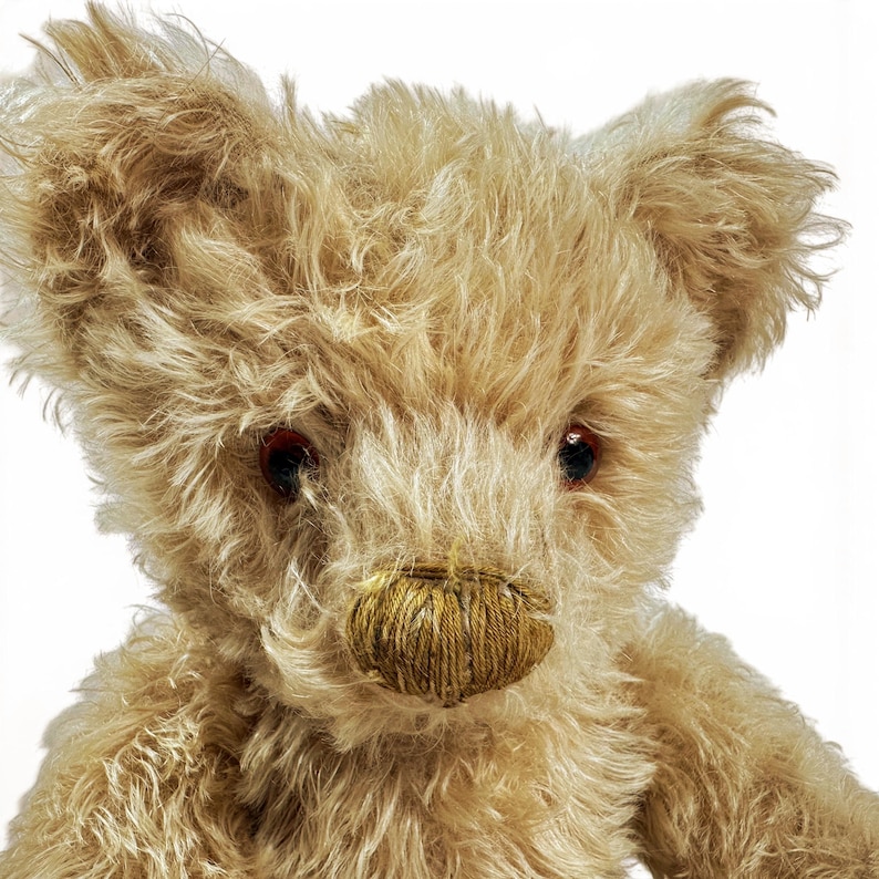 K&ouml;nnte beinhalten: Ein Pl&uuml;schteddyb&auml;r mit zotteligem, hellbraunem Fell, dunklen, runden, glasartigen Augen und einer gestickten, dunkelgoldbraunen Nase. Der B&auml;r hat ausgepr&auml;gte, abgerundete Ohren und ein strukturiertes, flauschiges Erscheinungsbild an Kopf und Schultern.