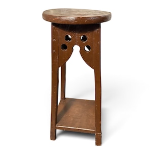 May include: A brown wooden plant stand featuring a round top, four vertical legs, and a lower square shelf. The upper section of the legs includes decorative cutouts with circular holes and arched supports, secured with visible metal nails.