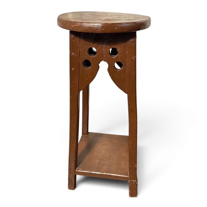 May include: A brown wooden plant stand featuring a round top, four vertical legs, and a lower square shelf. The upper section of the legs includes decorative cutouts with circular holes and arched supports, secured with visible metal nails.