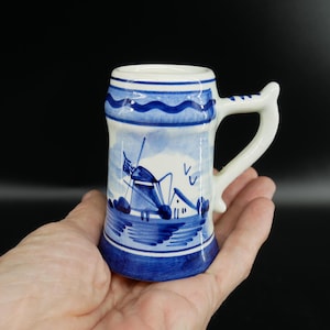 Könnte beinhalten: Ein kleiner Keramikbecher mit weißem Henkel und blau-weißem Windmühlendesign. Der Becher hat einen blauen Boden und Rand mit einer detaillierten Illustration einer Windmühle und einer Wasserszene. Der Becher wird in einer Hand gehalten.