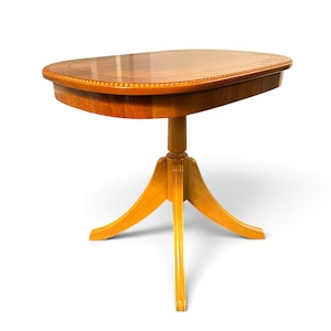 Op de afbeelding: Een houten zuiltafel met een ovaal blad met een gladde, gepolijste afwerking en een decoratieve kralenrand. De tafel wordt ondersteund door een centrale kolom die zich vertakt in drie gebogen poten voor een stabiele basis.