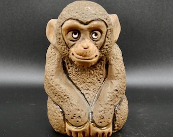 May include: A small, textured brown monkey figurine with a smooth face, large ears, and a seated posture. The sculpture features a detailed, porous surface texture across the body, contrasting with the defined, smooth facial features.