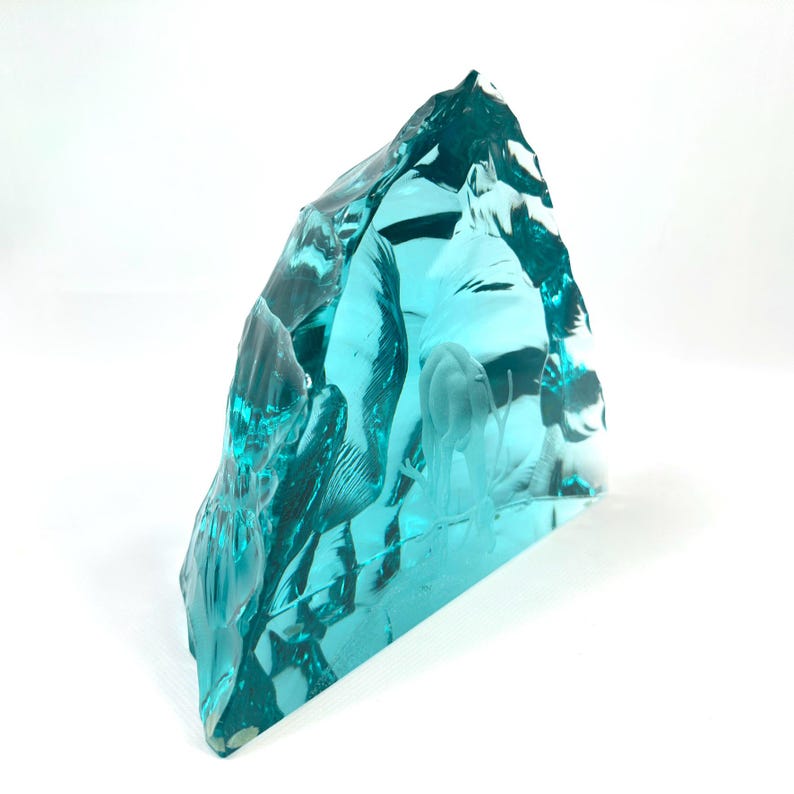 May include: A turquoise glass sculpture with a rough, textured surface. The sculpture is triangular in shape and features an etched design of what appears to be a jellyfish. The glass is translucent, allowing light to pass through.