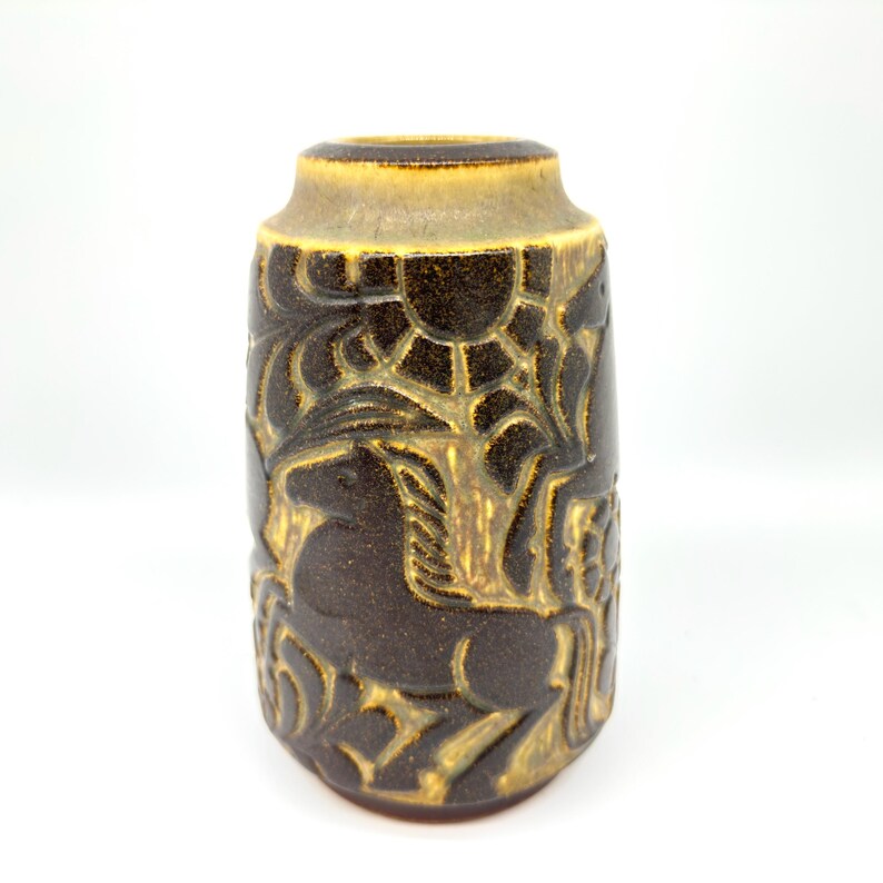 Peut inclure: Un vase en c&eacute;ramique marron de forme cylindrique avec un dessus l&eacute;g&egrave;rement &eacute;vas&eacute;. Le vase pr&eacute;sente un motif en relief d'un cheval, d'arbres et de soleil, avec des accents dor&eacute;s. Le vase mesure environ 15 cm de haut.