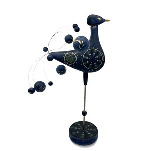 May include: A dark blue bird figurine standing on a thin metal rod and a circular base. The bird features a long tail made of thin wires tipped with small dark blue spheres. The body is decorated with green and white circular floral patterns.