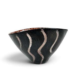 May include: A conical bowl with a textured black exterior featuring vertical, wavy silver brushstroke patterns. The interior of the bowl displays a contrasting metallic copper finish with visible linear markings along the upper rim.