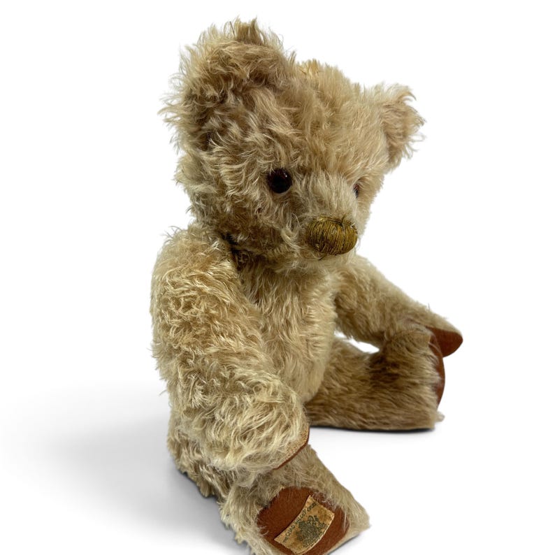 K&ouml;nnte beinhalten: Ein beigefarbener Teddyb&auml;r mit zotteligem Fell, dunkelbraunen Augen und einer gestickten Nase. Der B&auml;r hat braune Stoffpfoten, wobei an der Unterseite des rechten Fu&szlig;es ein kleines rechteckiges Etikett mit sichtbarem Text und Wappenmotiv angebracht ist.