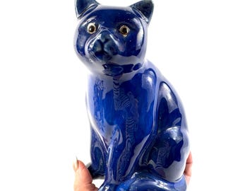 May include: A glossy, deep cobalt blue ceramic cat figurine in a seated position. The cat features a smooth, reflective surface, pointed ears, and round, light-coloured eyes with dark pupils, capturing a realistic feline form in a solid blue glaze.