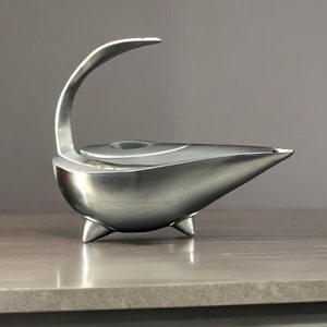 May include: A silver-toned metal oil lamp with a smooth, curved body, a pointed spout, and a high, arched handle. The vessel features a circular lid centered on top and is supported by three small, conical feet, presenting a sleek, polished finish.