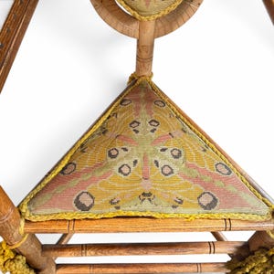 May include: A wooden chair featuring a triangular seat upholstered in fabric with a moth wing pattern in shades of yellow, green, and pink. The seat is edged with yellow braided trim, and the chair frame is constructed from turned wood.