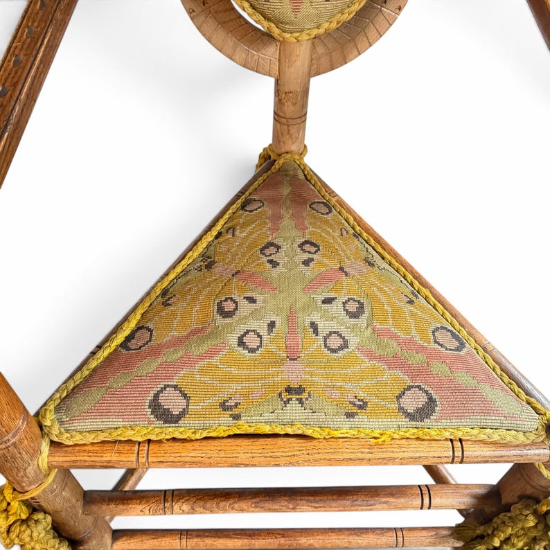 May include: A wooden chair featuring a triangular seat upholstered in fabric with a moth wing pattern in shades of yellow, green, and pink. The seat is edged with yellow braided trim, and the chair frame is constructed from turned wood.