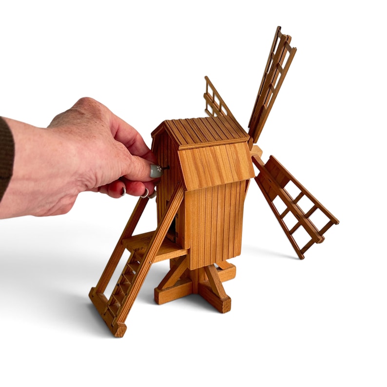 May include: A miniature wooden windmill model featuring vertical plank siding, a slanted roof, a set of four lattice-style blades, and an attached staircase leading to a small doorway. The structure is crafted from light brown wood with visible grain.