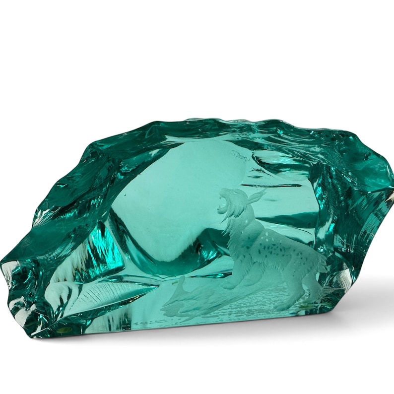 Peut inclure: Une sculpture en verre bleu sarcelle translucide pr&eacute;sentant une gravure d&eacute;taill&eacute;e d'un lynx debout dans un paysage rocheux. Le verre poss&egrave;de un bord ext&eacute;rieur irr&eacute;gulier et dentel&eacute; qui imite l'apparence d'une formation cristalline brute.
