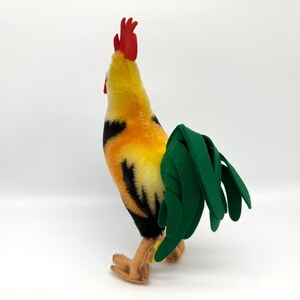 May include: A plush rooster toy with a vibrant color scheme. The rooster features a red comb, yellow and orange body, black accents, and a green tail. The toy is standing on a white surface.