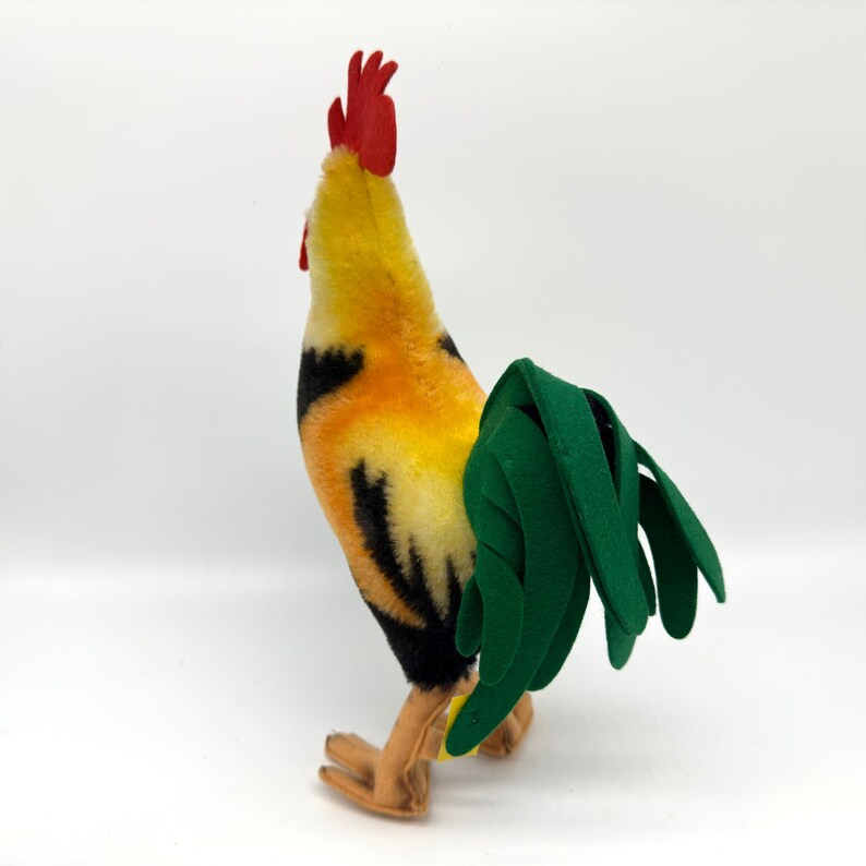 May include: A plush rooster toy with a vibrant color scheme. The rooster features a red comb, yellow and orange body, black accents, and a green tail. The toy is standing on a white surface.