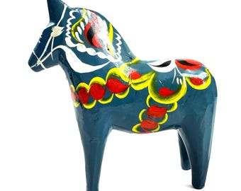 Puede incluir: Una figura de caballo de madera azul con patrones decorativos pintados a mano en blanco, amarillo y rojo. El caballo presenta un diseño estilizado de crines y silla de montar con líneas curvas y motivos florales en el cuerpo y las patas.