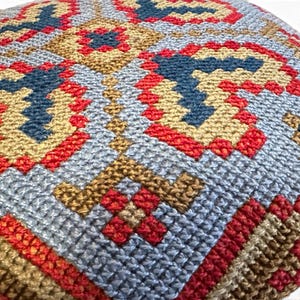 May include: A square cushion cover featuring a dense, hand-stitched needlepoint pattern. The design consists of geometric shapes in shades of light blue, deep red, and golden tan, creating a repeating, symmetrical, and intricate textured surface.