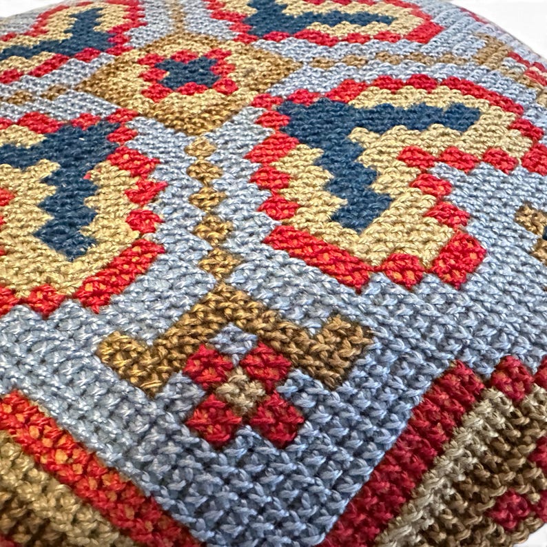 May include: A square cushion cover featuring a dense, hand-stitched needlepoint pattern. The design consists of geometric shapes in shades of light blue, deep red, and golden tan, creating a repeating, symmetrical, and intricate textured surface.