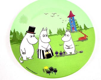 May include: A green ceramic plate featuring a whimsical illustration of Moomin characters in a landscape. The design includes three white, hippo-like figures, a red-roofed blue house, trees, and birds. The plate has a glossy finish.