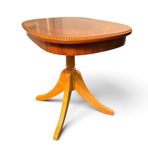 Op de afbeelding: Een houten bijzettafel met een glad, gepolijst ovaal blad met een donkere ingelegde rand en een kralenafwerking. De tafel wordt ondersteund door een centrale kolom die uitloopt in drie gebogen poten, uitgevoerd in een warme, lichtbruine houtnerf.