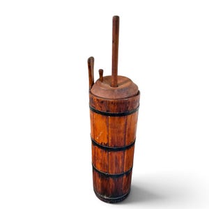 May include: A tall, cylindrical wooden butter churn featuring vertical staves held together by three dark, horizontal metal bands. It includes a circular wooden lid with a central vertical plunger handle and a small side handle attached to the top.