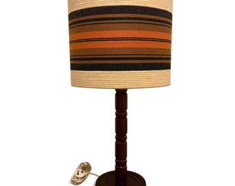 May include: A table lamp with a dark brown wooden base and a cylindrical shade. The shade features horizontal stripes in cream, black, dark brown, and orange. A white electrical cord with a two-prong plug extends from the base of the lamp.