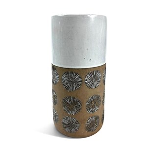 Könnte beinhalten: Eine zylindrische Keramikvase mit einem glatten, glänzend weißen oberen Teil und einem matten, hellbraunen unteren Teil. Der untere Bereich ist mit einem sich wiederholenden Muster aus dunklen, strukturierten, kreisförmigen Sternmotiven verziert.