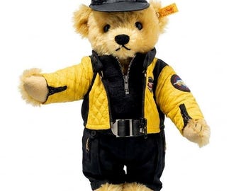 Puede incluir: Un oso de peluche con una chaqueta acolchada negra y amarilla, pantalones negros y una gorra negra. La chaqueta tiene una cremallera y un cinturón. La gorra tiene un logotipo en la parte delantera. El oso tiene una etiqueta en la oreja.