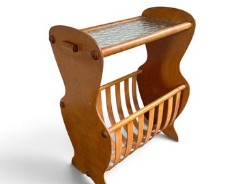 Peut inclure: Un porte-revues en bois brun clair doté d'une étagère supérieure avec un insert en verre translucide texturé. La structure se compose de deux panneaux latéraux incurvés reliés par une série de lattes en bois verticales formant un berceau.
