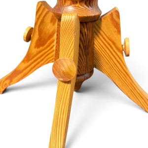 May include: A light brown wooden Christmas tree stand featuring a central cylindrical post with three extending legs. Each leg is secured with a round wooden knob, showcasing a natural wood grain pattern throughout the entire structure.