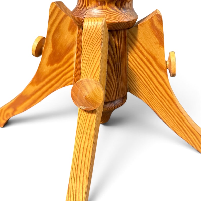 May include: A light brown wooden Christmas tree stand featuring a central cylindrical post with three extending legs. Each leg is secured with a round wooden knob, showcasing a natural wood grain pattern throughout the entire structure.