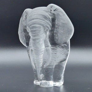 May include: A clear glass elephant sculpture. The elephant is standing with its trunk down and its ears spread. The sculpture is detailed, showing the wrinkles of the elephant's skin. The sculpture is on a dark background.
