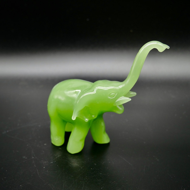 K&ouml;nnte beinhalten: Eine kleine, hellgr&uuml;ne Elefantenfigur mit erhobenem R&uuml;ssel. Die Figur ist glatt und gl&auml;nzend, mit sichtbaren Details der Ohren, Augen und Sto&szlig;z&auml;hne. Der Elefant steht auf einer dunklen Oberfl&auml;che.