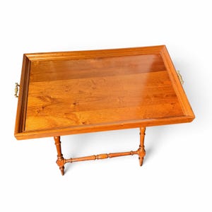 Puede incluir: Una mesa bandeja rectangular de madera con acabado pulido y veta visible. La bandeja tiene un borde elevado y peque&ntilde;as asas de lat&oacute;n en los laterales. Se apoya sobre cuatro patas de madera torneadas y delgadas con un travesa&ntilde;o.