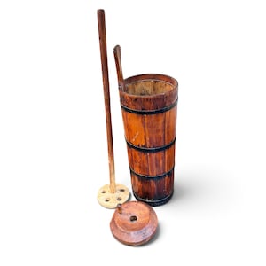 May include: A tall cylindrical wooden butter churn with dark metal bands, a long wooden dasher featuring a perforated circular base, and a separate circular wooden lid with a central hole, all showing natural wood grain and aged textures.