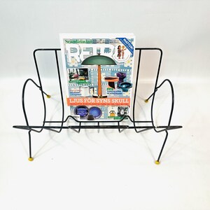 May include: A black metal magazine rack with a mid-century modern design. The rack holds a magazine with the text "Scandinavian Retro" and other text. The rack has yellow accents on the feet.