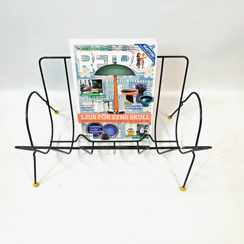 May include: A black metal magazine rack with a mid-century modern design. The rack holds a magazine with the text "Scandinavian Retro" and other text. The rack has yellow accents on the feet.