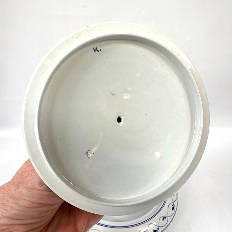 May include: A white ceramic cake stand lid with a blue floral design around the edge. The lid has a circular shape with a small hole in the centre and a blue marking on the top.