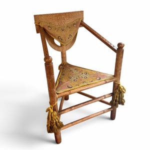 May include: A wooden chair with a triangular seat and backrest, both upholstered in a patterned fabric featuring moth-like motifs. The frame has carved geometric details, turned arm supports, and decorative gold-toned tassels on the front legs.