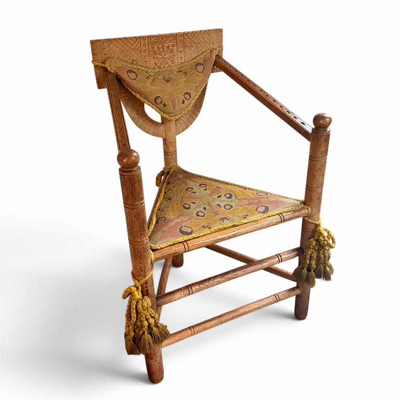 May include: A wooden chair with a triangular seat and backrest, both upholstered in a patterned fabric featuring moth-like motifs. The frame has carved geometric details, turned arm supports, and decorative gold-toned tassels on the front legs.