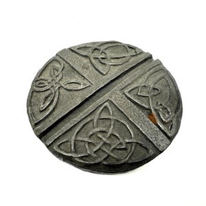 Op de afbeelding: Een rond, verweerd metalen object met een Keltisch knoopontwerp. Het object is donkergrijs van kleur en heeft een kruispatroon met ingewikkeld knoopwerk in elk kwadrant. Het ontwerp is verhoogd, waardoor het een driedimensionale uitstraling heeft.